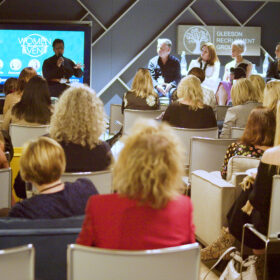 A photo of the Women in Leadership event. A crowd of people are facing a panel of talkers. There is a screen in the background with topics about leadership on.