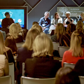 A photo of the Women in Leadership event, with the audience looking at the four pane speakers and the event presenter. The panel speakers are discussing key topics, while the audience look on.