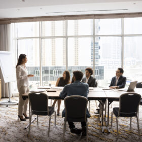 A boardroom meeting with professionals. A woman is standing at the head of the table, presenting ideas to the group.