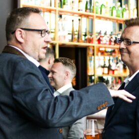 Two business men holding an indepth conversation at a networking event. They are both dressed in suits and are looking at each other, engaged in conversation. They are in a bar setting, and there are other professionals networking in the background of the shot.