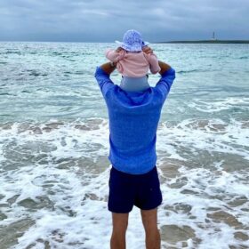 A photo of the author, Andrew, with his young daughter. They are stood in front of the sea facing the water, and his daughter is sat on his shoulders.